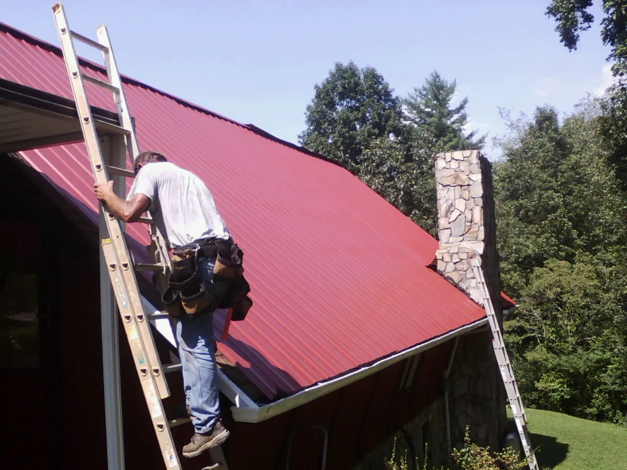 Roofer on a ladder beside a red metal roof and stone chimney — Soffit & Fascia Repair by Roofing Brothers in Satartia
